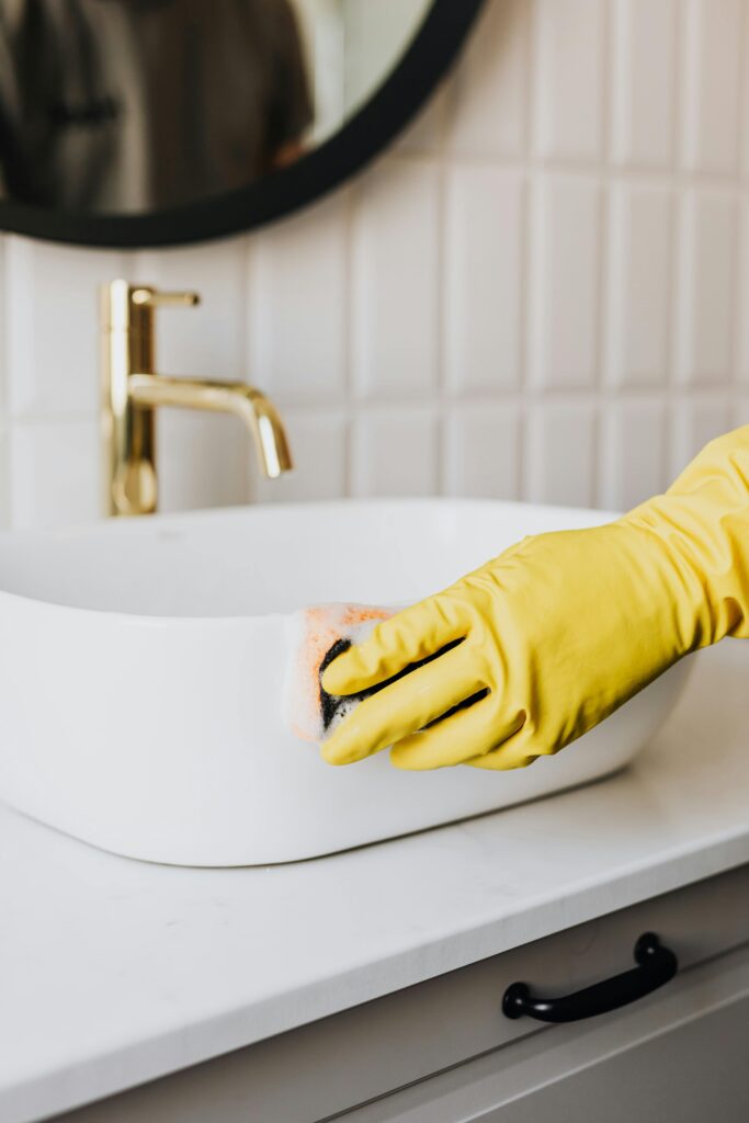 pexels-photo-4239110-4239110 Crop faceless person in yellow rubber protective glove wiping surface of white sink in bathroom with sponge and cleaning agent